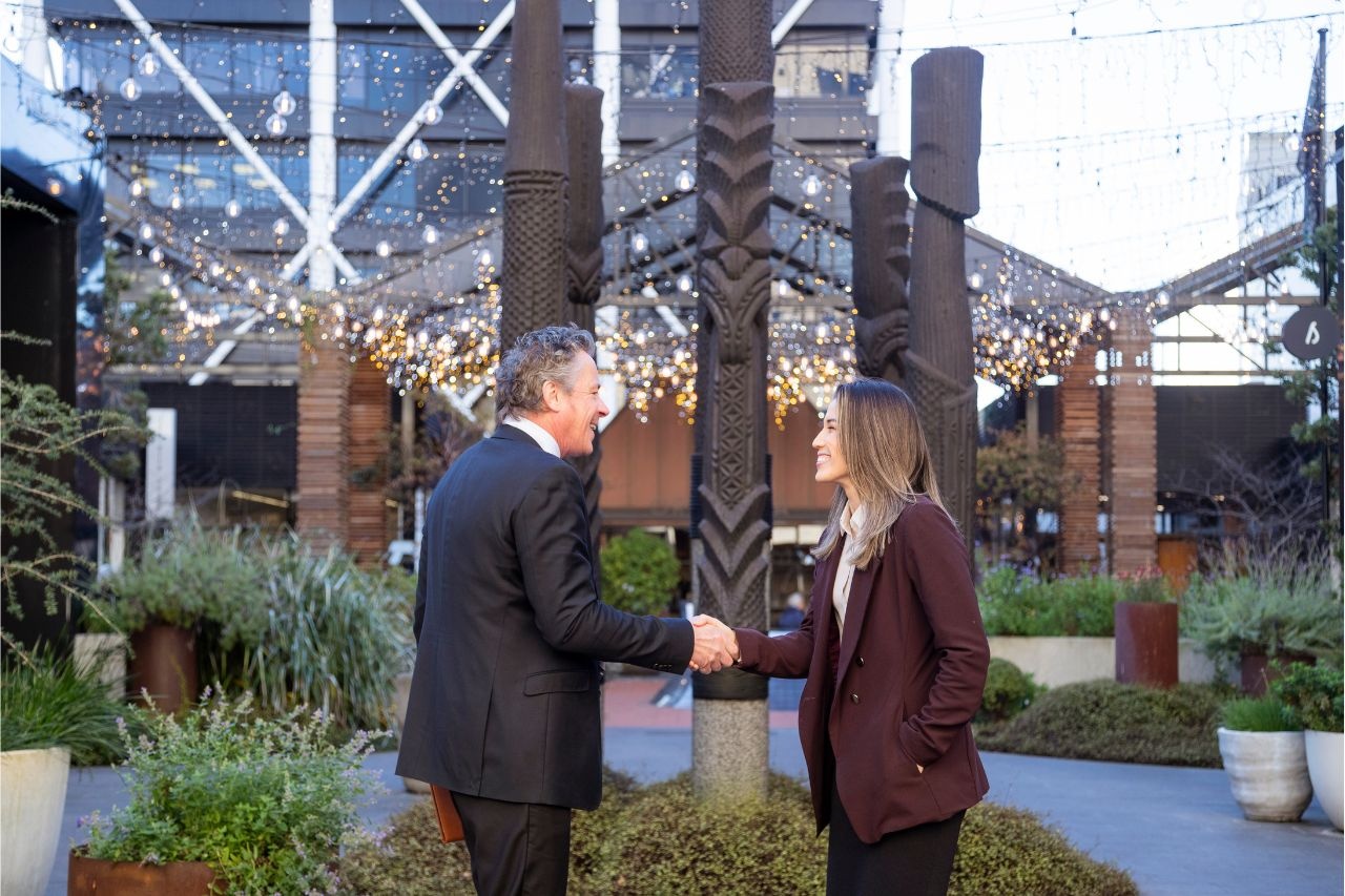 Auckland Business Events Fund Gallery Image 4 Two business professionals shaking hands in an outdoor Auckland venue featuring Māori carvings, symbolising partnerships supported by the Auckland Business Events Fund.